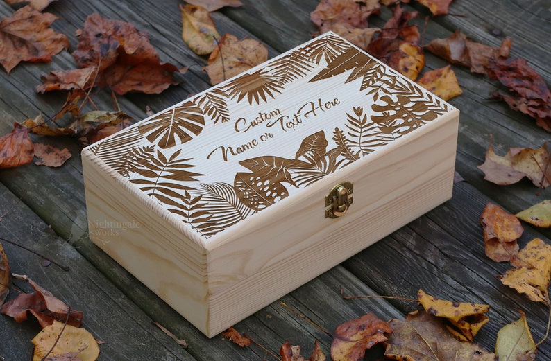 May include: A wooden box with a hinged lid and a brass latch. The lid is decorated with a laser-cut design of tropical leaves and the text "Custom Name or Text Here".