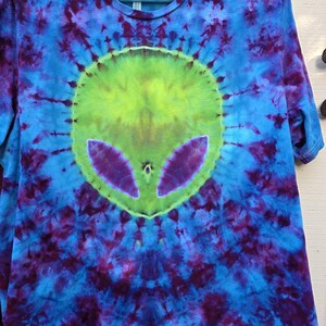 Pode incluir: Camiseta tie-dye com um design de rosto alien&iacute;gena. A camisa apresenta um esquema de cores vibrantes de azul, roxo e verde. O rosto do alien&iacute;gena est&aacute; centrado na camisa, com olhos grandes e uma forma de cabe&ccedil;a distinta.