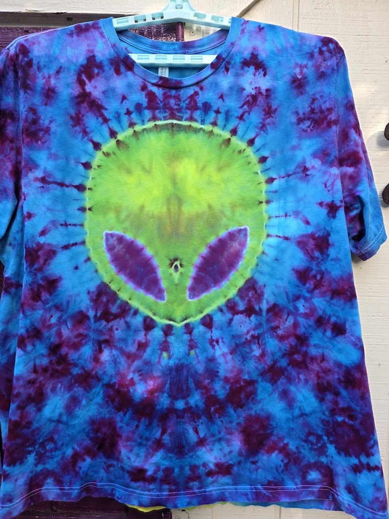 Pode incluir: Camiseta tie-dye com um design de rosto alien&iacute;gena. A camisa apresenta um esquema de cores vibrantes de azul, roxo e verde. O rosto do alien&iacute;gena est&aacute; centrado na camisa, com olhos grandes e uma forma de cabe&ccedil;a distinta.