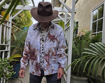 May include: A brown fedora hat paired with a light blue button-down shirt featuring a unique tie-dye pattern in shades of brown and red. The shirt is worn over a patterned top, and the person is wearing dark gray pants.