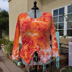 May include: An orange, red, and white tie-dye long-sleeve shirt with a scoop neck and a drawstring closure. The shirt has a floral design on the front and a scalloped hemline. The shirt is displayed on a mannequin.
