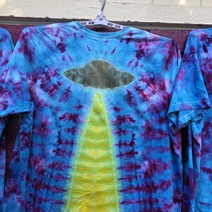 Pode incluir: Camiseta tie-dye de manga comprida com um design azul, roxo e amarelo. A camisa apresenta um gr&aacute;fico de OVNI cinza com um feixe de luz amarelo. A camisa est&aacute; pendurada em um cabide branco.