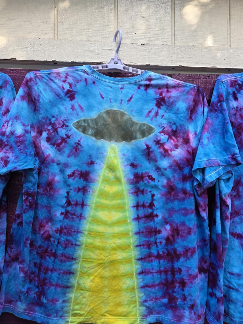 Pode incluir: Camiseta tie-dye de manga comprida com um design azul, roxo e amarelo. A camisa apresenta um gr&aacute;fico de OVNI cinza com um feixe de luz amarelo. A camisa est&aacute; pendurada em um cabide branco.
