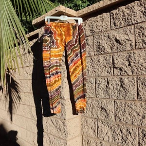 May include: Tie-dye leggings in shades of orange, brown, and purple hang on a white hanger against a textured brick wall. The leggings feature a striped pattern with varying widths and colors, creating a unique design. The garment is made of a soft, lightweight material.