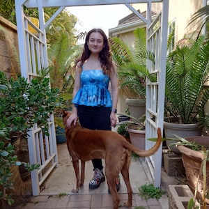 May include: A woman wearing a blue tie-dye top and black jeans stands with a brown dog in a garden setting. The woman is smiling and petting the dog. The dog is looking at the camera.