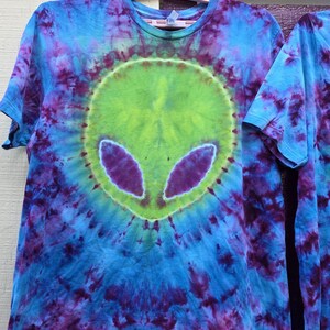 Pode incluir: Camiseta tie-dye com um design de rosto alien&iacute;gena. A camisa &eacute; predominantemente azul, com detalhes roxos e verdes. O rosto alien&iacute;gena &eacute; verde com olhos roxos, em um fundo tie-dye em espiral.