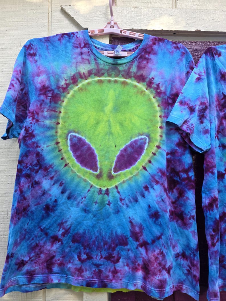 Pode incluir: Camiseta tie-dye com um design de rosto alien&iacute;gena. A camisa &eacute; predominantemente azul, com detalhes roxos e verdes. O rosto alien&iacute;gena &eacute; verde com olhos roxos, em um fundo tie-dye em espiral.