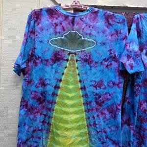 Pode incluir: Camiseta tie-dye com um design azul, roxo e amarelo. A camisa apresenta um gr&aacute;fico de nuvem e feixe. A camisa de manga curta est&aacute; pendurada em um cabide rosa em um fundo branco e roxo.