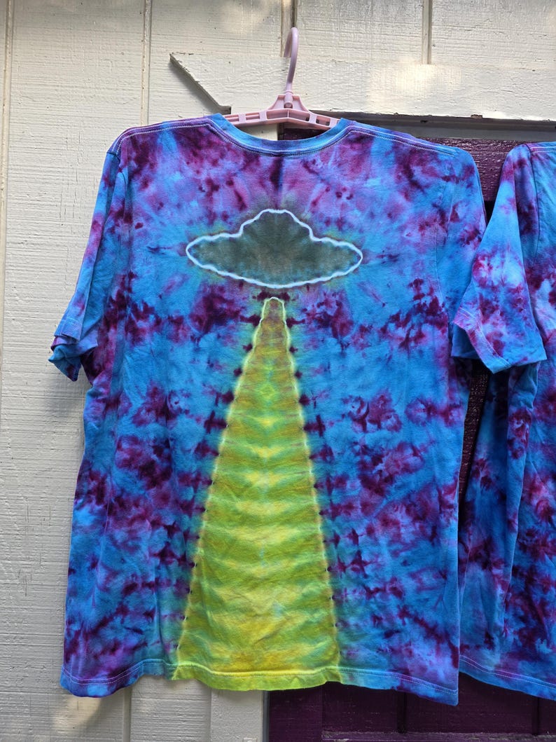 Pode incluir: Camiseta tie-dye com um design azul, roxo e amarelo. A camisa apresenta um gr&aacute;fico de nuvem e feixe. A camisa de manga curta est&aacute; pendurada em um cabide rosa em um fundo branco e roxo.