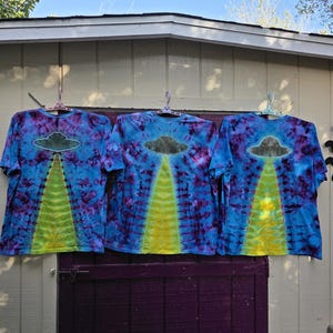 Pode incluir: Tr&ecirc;s camisetas tie-dye em azul, roxo e amarelo. Cada camisa apresenta um gr&aacute;fico de um OVNI com um raio de luz amarelo. As camisas est&atilde;o penduradas em cabides rosa. Fundo branco e roxo.