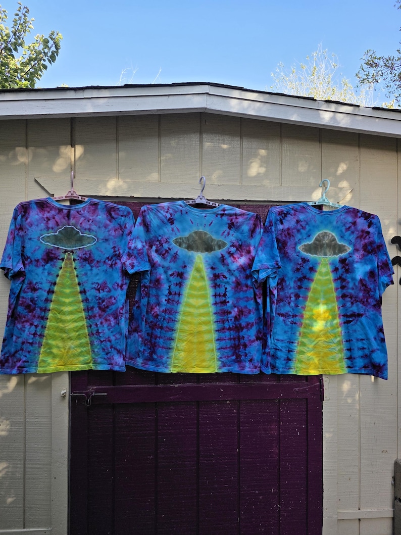 Pode incluir: Tr&ecirc;s camisetas tie-dye em azul, roxo e amarelo. Cada camisa apresenta um gr&aacute;fico de um OVNI com um raio de luz amarelo. As camisas est&atilde;o penduradas em cabides rosa. Fundo branco e roxo.