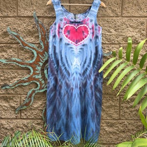 May include: A sleeveless tie-dye dress in shades of blue and purple, with a large pink heart design on the chest. The dress is hanging on a wooden hanger against a textured brick wall.