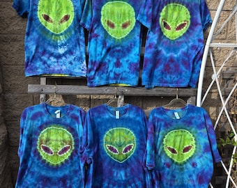 Peut inclure: Six t-shirts à manches courtes présentant un motif tie-dye dans des tons de bleu et de violet. Chaque t-shirt affiche un motif circulaire central représentant une tête d'extraterrestre vert vif aux yeux sombres en amande.