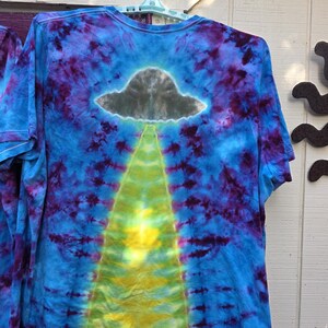 Pode incluir: Camiseta tie-dye com um design azul, roxo e amarelo. A camiseta apresenta um gr&aacute;fico de OVNI cinza com um raio de luz amarelo. A camiseta est&aacute; pendurada em um cabide azul claro.