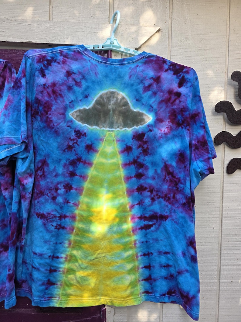 Pode incluir: Camiseta tie-dye com um design azul, roxo e amarelo. A camiseta apresenta um gr&aacute;fico de OVNI cinza com um raio de luz amarelo. A camiseta est&aacute; pendurada em um cabide azul claro.
