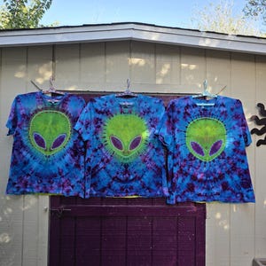 Pode incluir: Tr&ecirc;s camisetas tie-dye com um design de rosto alien&iacute;gena. As camisas s&atilde;o azuis com detalhes roxos e verdes. Cada camisa tem um rosto alien&iacute;gena verde com olhos roxos no centro. As camisas est&atilde;o penduradas em um varal contra um fundo branco e roxo.