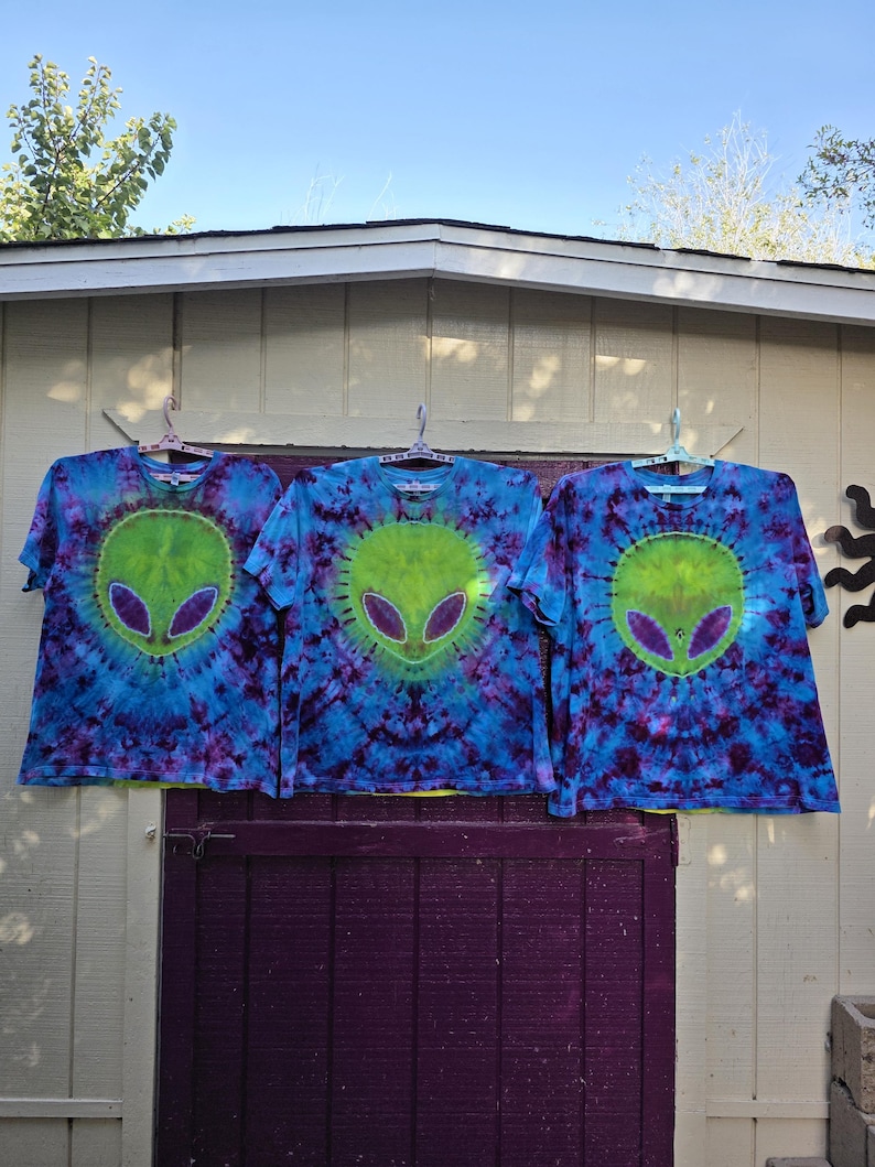 Pode incluir: Tr&ecirc;s camisetas tie-dye com um design de rosto alien&iacute;gena. As camisas s&atilde;o azuis com detalhes roxos e verdes. Cada camisa tem um rosto alien&iacute;gena verde com olhos roxos no centro. As camisas est&atilde;o penduradas em um varal contra um fundo branco e roxo.