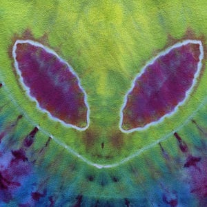 Pode incluir: Tecido tie-dye com um design de rosto alien&iacute;gena. O design apresenta dois grandes olhos ovais roxos delineados em branco, contra um fundo amarelo-esverdeado. A parte inferior transita para tons de azul e roxo.