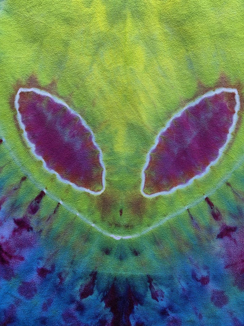 Pode incluir: Tecido tie-dye com um design de rosto alien&iacute;gena. O design apresenta dois grandes olhos ovais roxos delineados em branco, contra um fundo amarelo-esverdeado. A parte inferior transita para tons de azul e roxo.