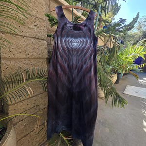 May include: A sleeveless, knee-length dress with a tie-dye pattern in shades of purple, black, and white. The dress is hanging on a wooden hanger outdoors, with a backdrop of greenery and a stone wall.