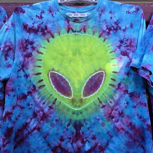 Pode incluir: Camiseta tie-dye com um design de rosto alien&iacute;gena. O rosto alien&iacute;gena &eacute; verde lim&atilde;o com olhos roxos, em um fundo tie-dye azul e roxo. A camiseta est&aacute; pendurada em um cabide branco.