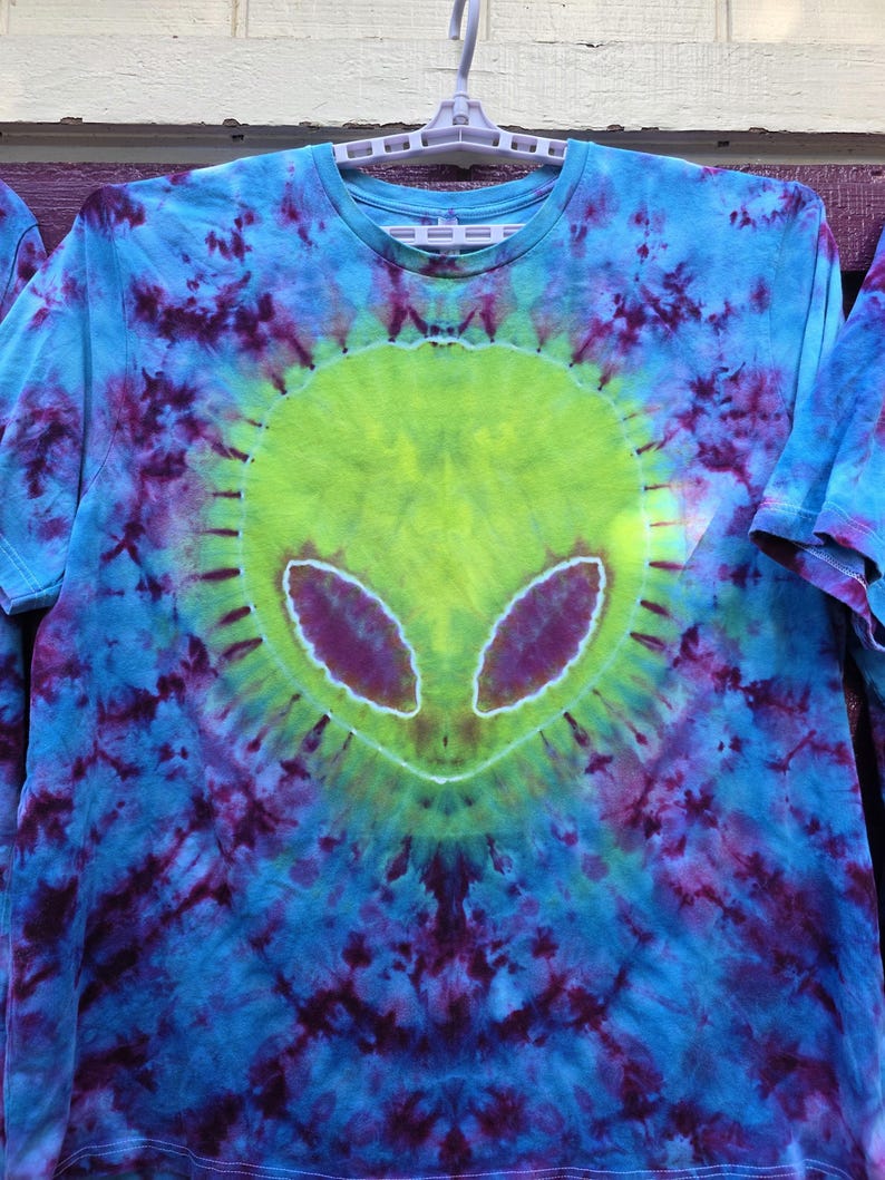 Pode incluir: Camiseta tie-dye com um design de rosto alien&iacute;gena. O rosto alien&iacute;gena &eacute; verde lim&atilde;o com olhos roxos, em um fundo tie-dye azul e roxo. A camiseta est&aacute; pendurada em um cabide branco.
