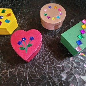 May include: Four small decorated boxes. A yellow hexagonal box with colorful gems, a red heart-shaped box with painted flowers, a brown round box with colorful stars, and a green rectangular box with colorful gems.