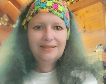 May include: A woman with teal hair is wearing a colorful crocheted headband with a button on the side.