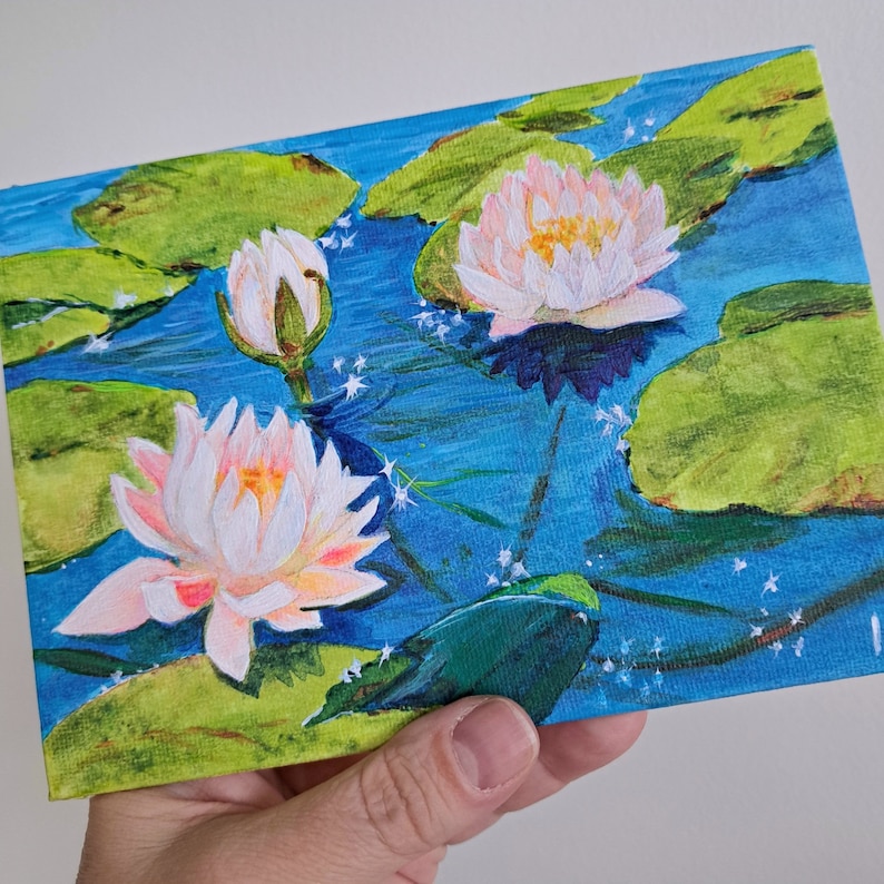 May include: A painting of white water lilies with pink centres on a blue background with green lily pads. The painting has a sparkling effect.