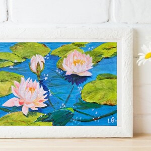May include: A painting of pink water lilies with green lily pads on a blue background. The painting is framed in white.