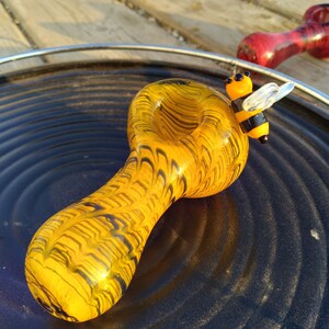 May include: A yellow and black glass pipe with a bee design on the side. The pipe is sitting on a black surface.