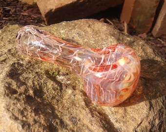 Op de afbeelding: Een glazen pijp met een helder lichaam en oranje en witte swirls. De pijp ligt op een rots.