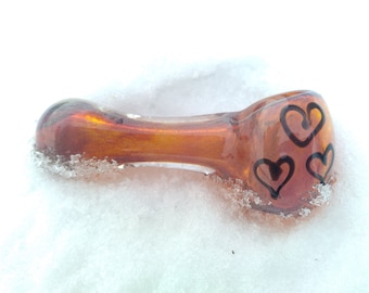 Op de afbeelding: Amberkleurige glazen pijp met drie zwarte harten aan de zijkant, liggend op sneeuw.