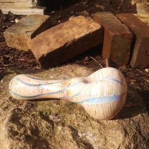 Pode incluir: Um cachimbo de vidro com um design marmorizado marrom claro e azul. O cachimbo está sentado em uma rocha grande, áspera e cinza.