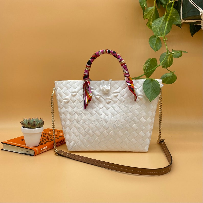 May include: White woven tote bag with a colorful patterned fabric handle and a gold chain strap. The bag is sitting on a light brown surface.