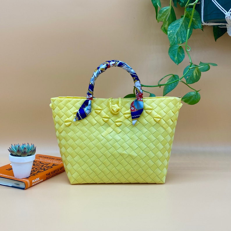 May include: A yellow woven tote bag with a blue and white patterned fabric handle. The bag is made of a woven material and has a rectangular shape.
