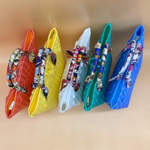 May include: Five colorful woven tote bags with different colored fabric handles. The bags are arranged in a row from left to right, with the red bag on the left and the blue bag on the right. The bags are all made of a woven material and have a unique design.