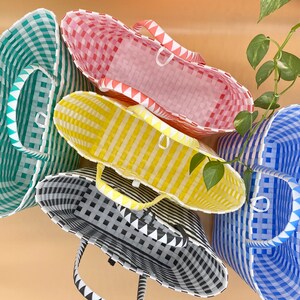 May include: Four woven plastic tote bags in different colors and patterns. The bags are stacked on top of each other. The top bag is pink and white, the second bag is yellow and white, the third bag is black and white, and the bottom bag is blue and white.