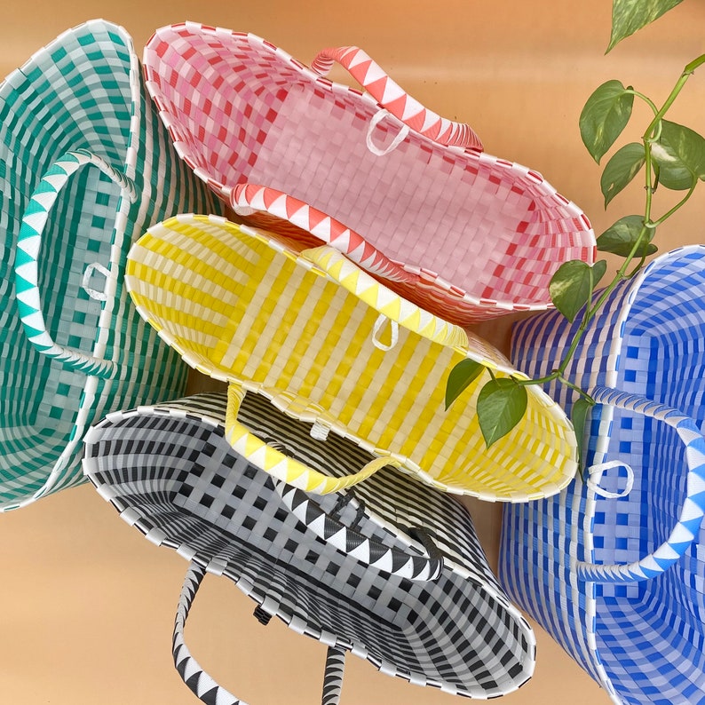 May include: Four woven plastic tote bags in different colors and patterns. The bags are stacked on top of each other. The top bag is pink and white, the second bag is yellow and white, the third bag is black and white, and the bottom bag is blue and white.