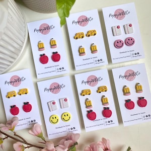 May include: A selection of handmade stud earrings displayed on white cards. The earrings showcase school-related designs, such as yellow school buses, pencils, red apples, notebooks, and smiley faces. The cards are labelled "Poppy Wild Co" and "Handmade in Ontario".