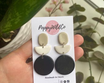 May include: A pair of handmade geometric earrings with black, white, and beige clay circles and half-circles. The earrings are on a white card with the text "Poppy WildCo" and "Handmade in Ontario".