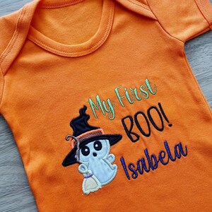 May include: Orange baby bodysuit with the embroidered text "My First BOO!" and the name "Isabela". The design includes a cartoon ghost wearing a witch hat and holding a broom, perfect for Halloween.