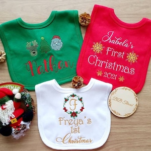 May include: Three baby bibs in green, red, and white, embroidered with Christmas-themed designs and names. The green bib features a reindeer, Christmas tree, and Santa. The red bib says "Isabela's First Christmas 2021". The white bib says "Freya's 1st Christmas".