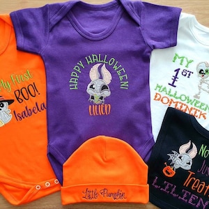 May include: A collection of Halloween-themed baby outfits in orange, purple, white, and black. The outfits include onesies and a beanie, each embroidered with spooky designs and text like "My First BOO!" and "Happy Halloween!"