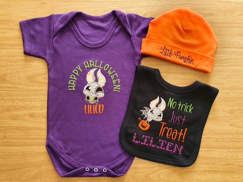 May include: A Halloween-themed baby outfit set featuring a purple onesie with "Happy Halloween!" and a bat design, an orange beanie with "Little Pumpkin", and a black bib with "No trick Just Treat!" and a bat design.