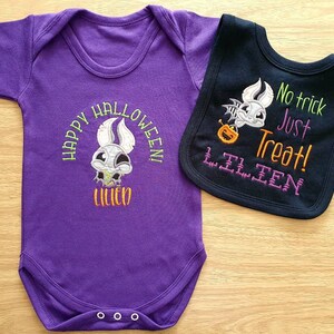 May include: A purple baby bodysuit and a black bib set. The bodysuit has a Halloween design with the text "HAPPY HALLOWEEN! LILIEN". The bib features a bat with a pumpkin and the text "No trick Just Treat! LILIEN".