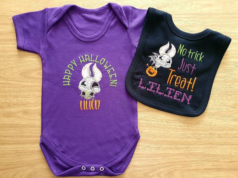 May include: A purple baby bodysuit and a black bib set. The bodysuit has a Halloween design with the text "HAPPY HALLOWEEN! LILIEN". The bib features a bat with a pumpkin and the text "No trick Just Treat! LILIEN".