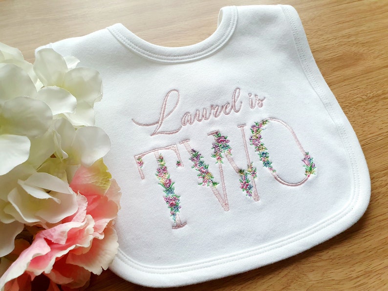 May include: White baby bib with embroidered text "Laurel is TWO". The word "TWO" is decorated with floral embroidery in pink, green, and yellow. The bib is surrounded by pink and white flowers.
