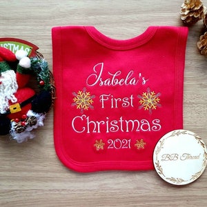 May include: A red baby bib embroidered with "Isabela's First Christmas 2021" and snowflake designs. A wooden disc with "BB Thread" is next to the bib. A Christmas wreath and pine cones are in the background.