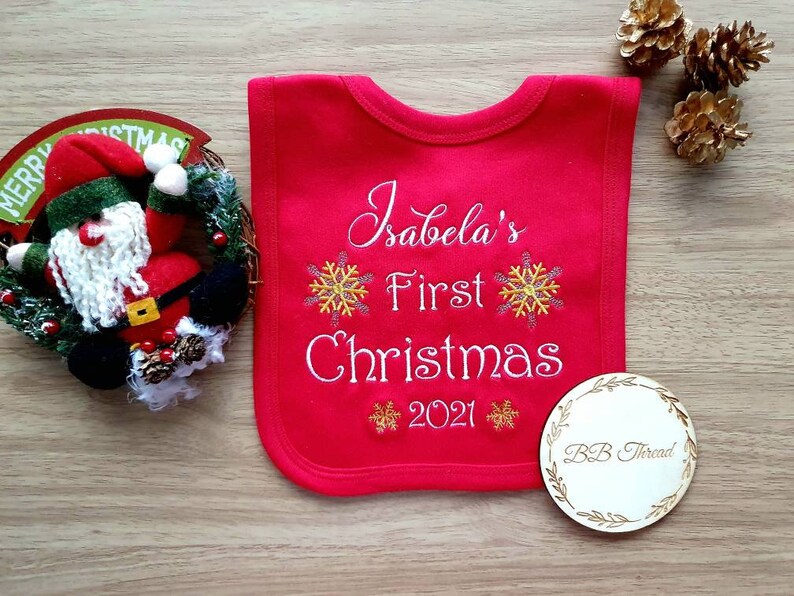 May include: A red baby bib embroidered with "Isabela's First Christmas 2021" and snowflake designs. A wooden disc with "BB Thread" is next to the bib. A Christmas wreath and pine cones are in the background.